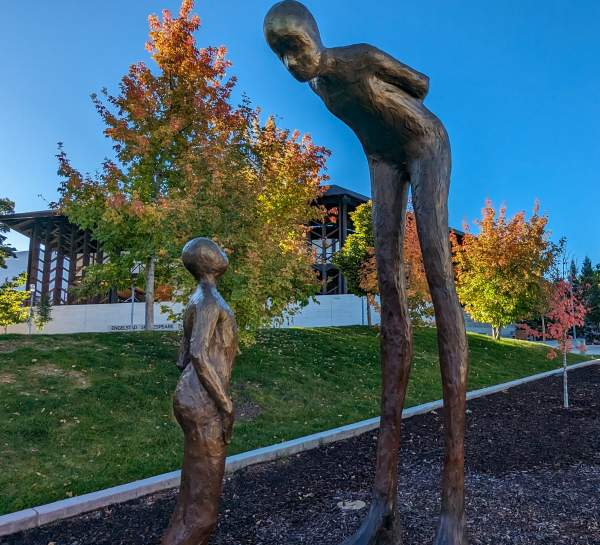 Two large bronze-cast sculptures stand opposite each other in front of a line of trees with vibrant fall leaves. The humanoid sculpture on the right is taller than the one on the left and looking down at them as if in conversation.