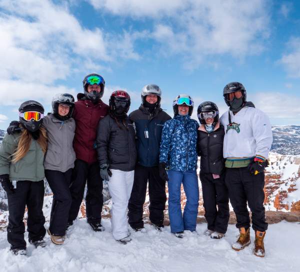 A group of eight adults dressed for winter recreation stand together for a photo at the North Overlook of Cedar Breaks National Monument