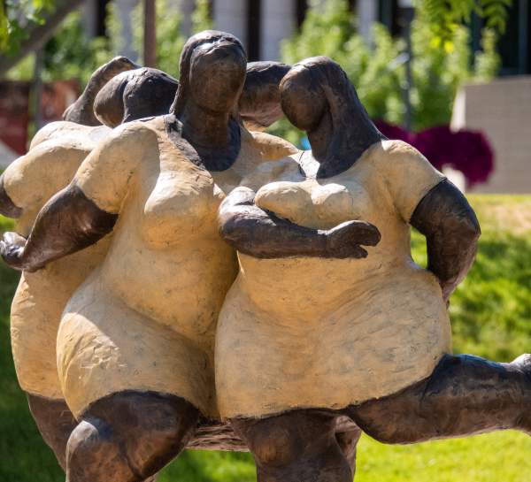 A dark metal sculpture of three women dancing together, back to back, shoulder to shoulder.
