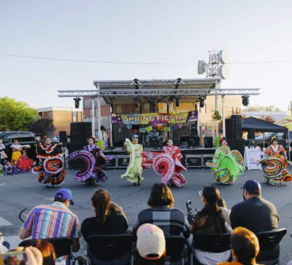 Traditional folklórico dancers performing at a Cedar City street festival with colorful dresses and live stage in background