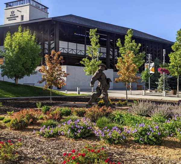 A bronze-cast sculpture of a man wearing a wide-brimmed hat scatters seeds along the ground in a flower garden in front of a large theater.