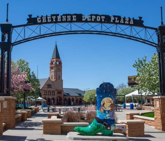 A colorful scene at Cheyenne Depot Plaza, where a painted cowboy boot stands in the middle of the courtyard as vibrant trees bloom in the background before big clock tower. 