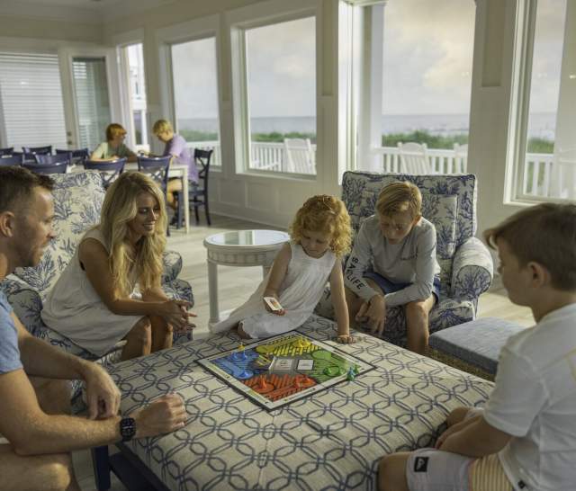A family sits together in a bright, coastal-style beach house playing a board game. Large windows behind them reveal an oceanfront view, and other guests are seen relaxing in the background, creating a warm and inviting atmosphere.