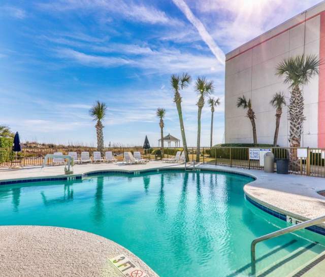 Outdoor swimming pool surrounded by lounge chairs and palm trees at a beachfront hotel. A clear blue sky and a pink-accented building are visible, with the beach and ocean just beyond the pool area.
