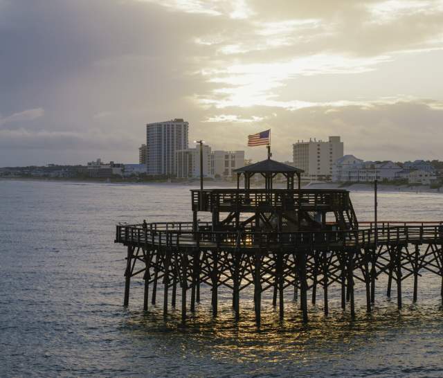 Cherry Grove Pier extends over the ocean in North Myrtle Beach. The American flag waves atop the pier’s iconic octagonal pavilion, and the sun reflects off the water below. A few people are seen fishing along the pier, while the shoreline in the background is lined with coastal homes and high-rise buildings.