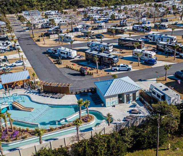 Aerial view of a large RV resort featuring rows of parked motorhomes and trailers. In the foreground, there’s a pool area with a waterslide, splash features, and a poolside building, surrounded by palm trees and landscaped areas