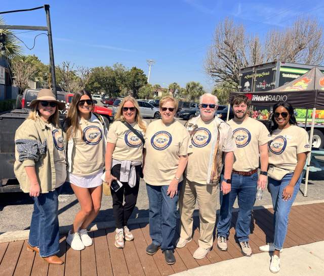Seven people wearing matching event T-shirts stand smiling outdoors in sunny weather. They pose together in front of vendor trucks and an iHeartRadio tent at the inaugural Oyster Roast for the Coast.