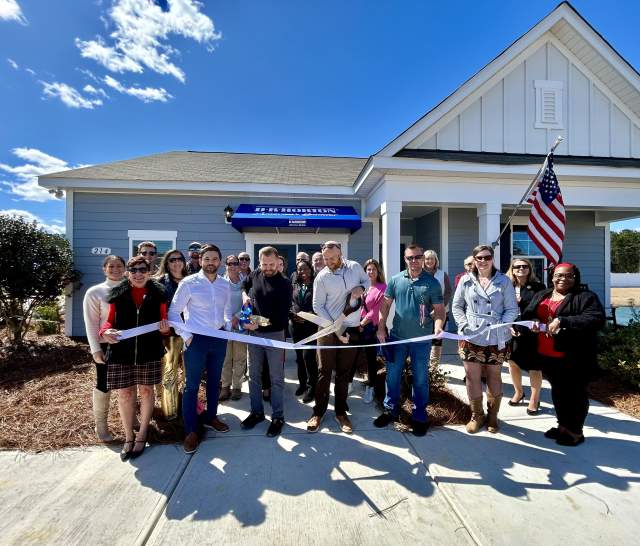 A large group of people stands in front of a light blue building with a D.R. Horton awning for a ribbon cutting ceremony. They hold a long white ribbon while one person uses oversized scissors. An American flag is displayed beside the building under a bright blue sky with scattered clouds.