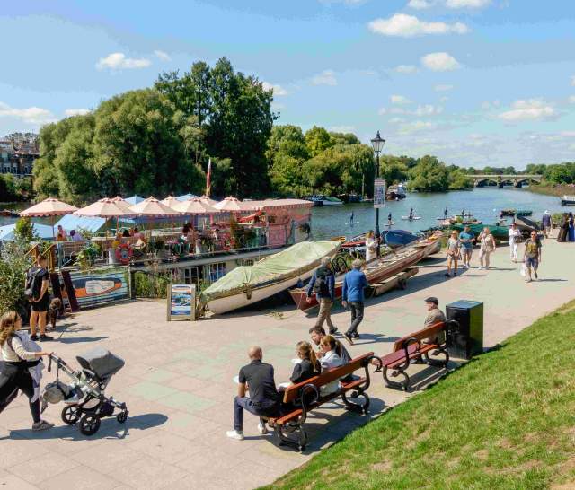 People sitting on benches or walking along the Thames Path at Richmond Upon Thames. Boats and river in the background.
