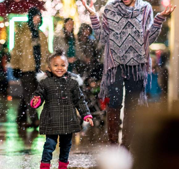 A mom and daughter with decorative holiday lights behind them look with anticipation toward a fancy chair