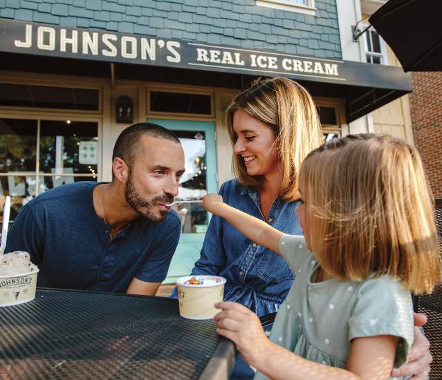 A young daughter feeding ice cream to her dad while her mom watches on the front patio of  Johnson's Ice Cream