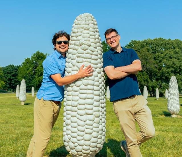 A couple of friends at the Field of Corn. One of them hugging a corn and the other leaning on a corn.