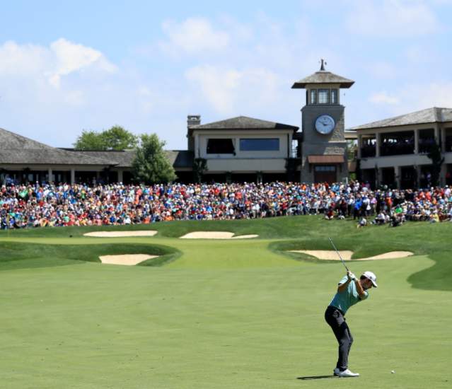 Golfer swining club at golf ball towards the 18th green that is surrounded by a crowd at the Memorial Touranemnt.
