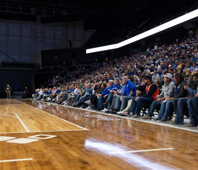 Large crowd seated courtside and in arena stands during a basketball game, with fans watching the action from rows along the hardwood floor under bright indoor lighting.