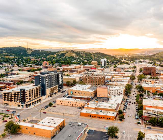 Aerial view of downtown Rapid City at sunset, with downtown streets lined by low-rise buildings, cars, and parking lots. Rolling green hills surround the city, while dramatic clouds stretch across the sky and golden sunlight glows on the horizon.