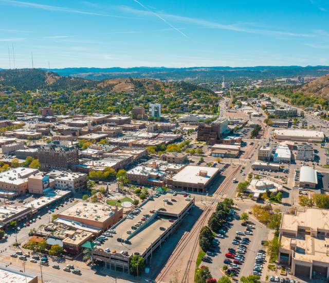 Aerial view of downtown Rapid City with buildings, roads, and parking lots. Surrounding green hills and a clear blue sky create a serene and expansive atmosphere.