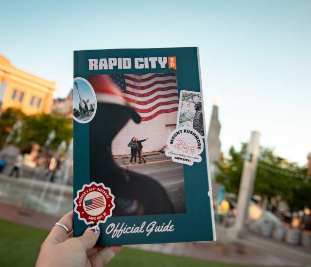 The cover of the Visit Rapid City guide is held up by a woman against the backdrop of the fountains at Main Street Square.