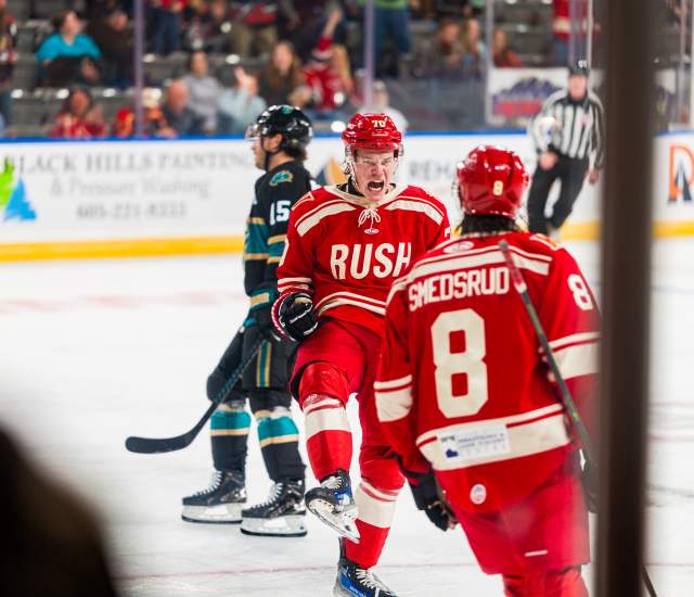 A player in a red Rush jersey yells after scoring a goal, skating towards a teammate in the foreground.