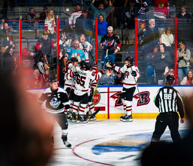 Hockey players in white jerseys gather for a group hug after scoring a goal. Fans behind the boards stand and celebrate with the team.