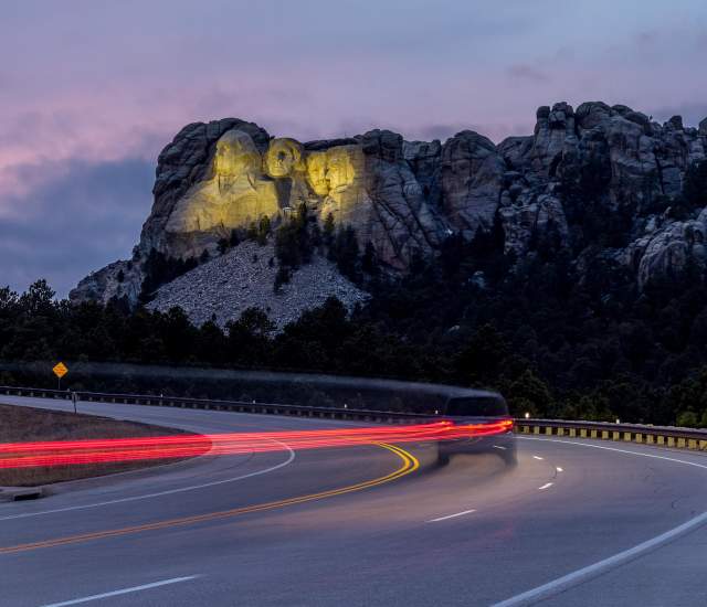 Mount Rushmore, lit up by yellow floodlights at dusk, sits in the background as a vehicle traveling down the highway is trailed by a long line of red taillights, created by a long-exposure photo.