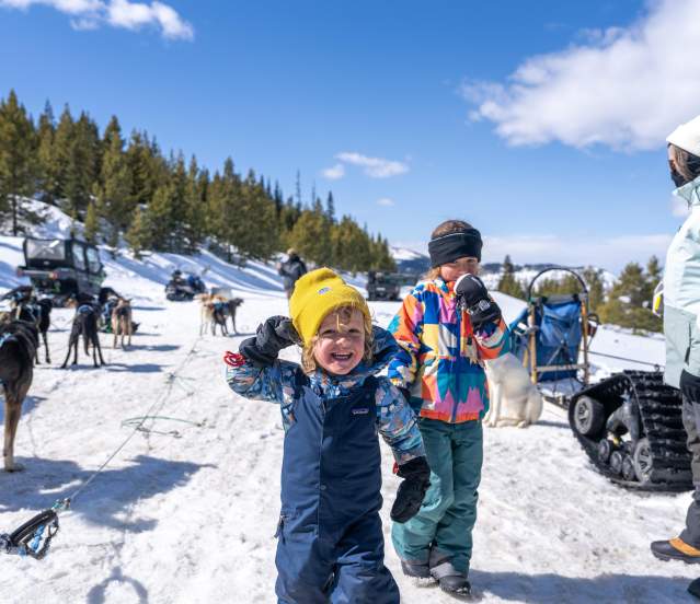 Two children play in a snowy landscape, surrounded by sled dogs and snow-covered trees. A person in winter gear stands nearby, with vehicles in the background.