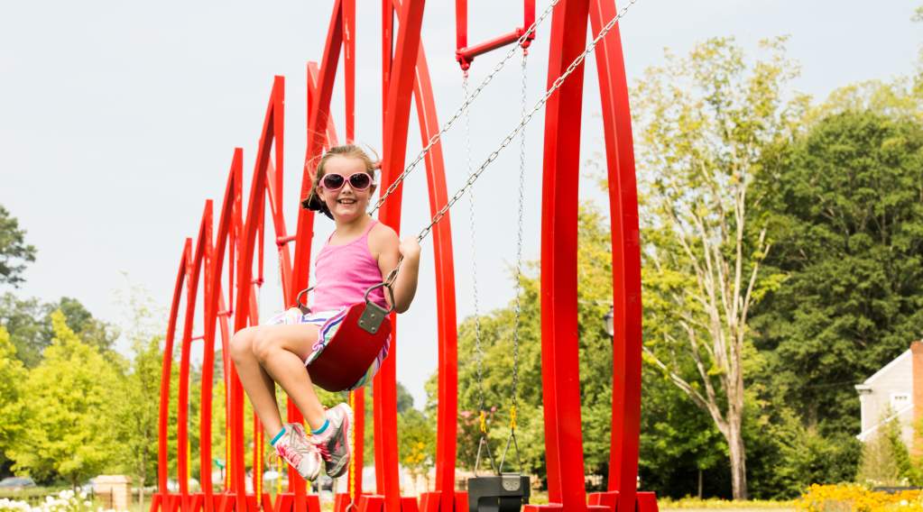 The red artistic swing set and lush trees at Abernathy Greenway Playable Art Park