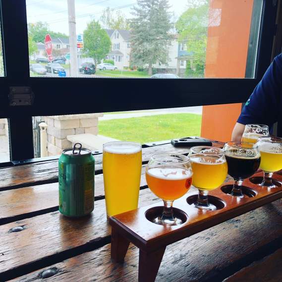a flight of beer lined up on a wooden bar looking out a window with greenery