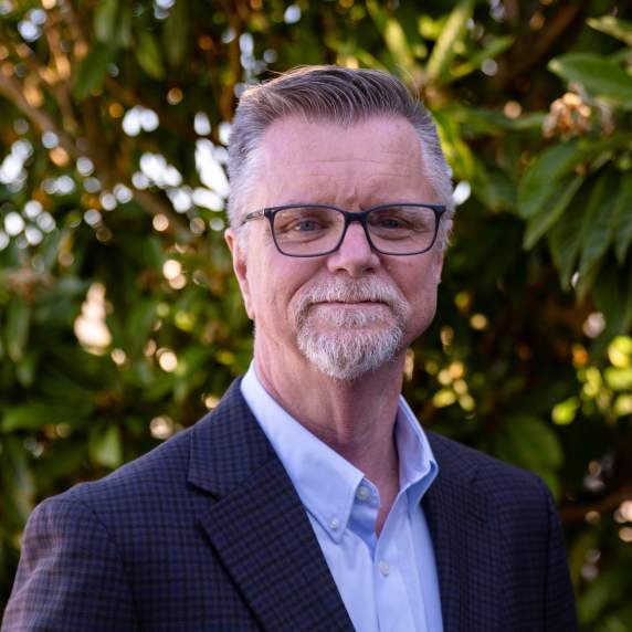 Outdoor headshot of a man with short gray hair, glasses, and a gray goatee wearing a dark checkered blazer and light blue button-down shirt, standing in front of leafy greenery.