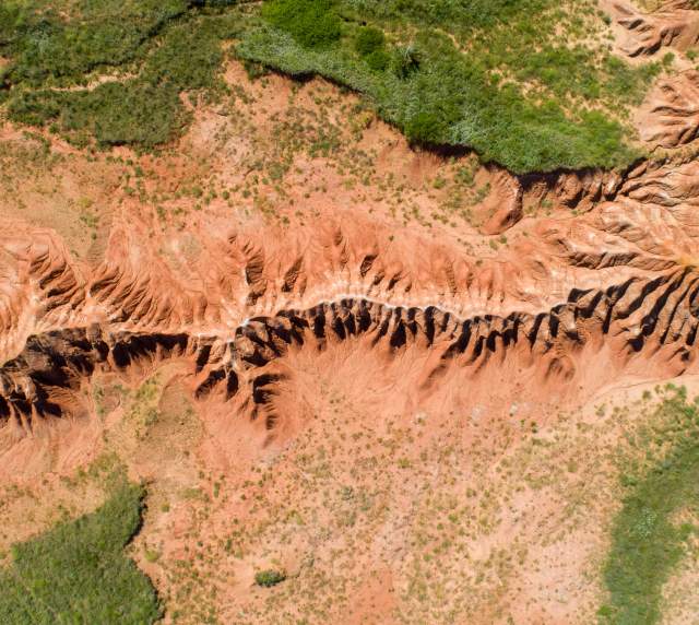 Looking down at a rugged landscape of red hills that looks like a spine. Patchy green grass surrounds the hills