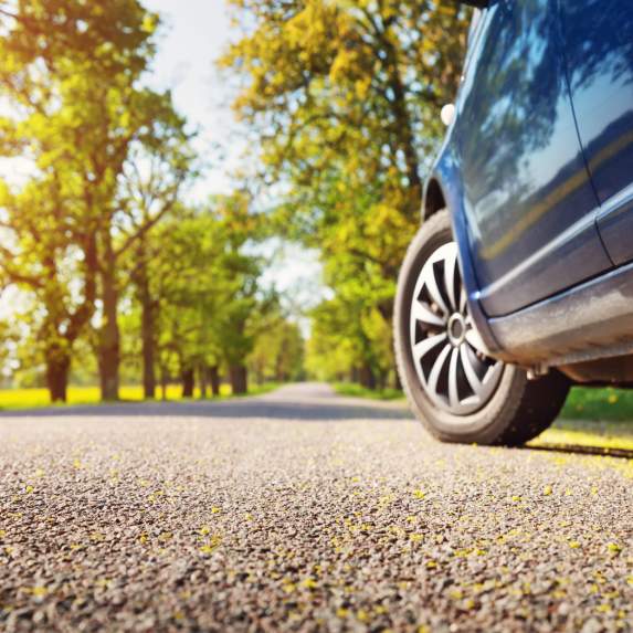 Car driving along a sunlit tree-lined road, featuring a close-up view of a vehicle wheel and scenic travel through a green outdoor landscape.