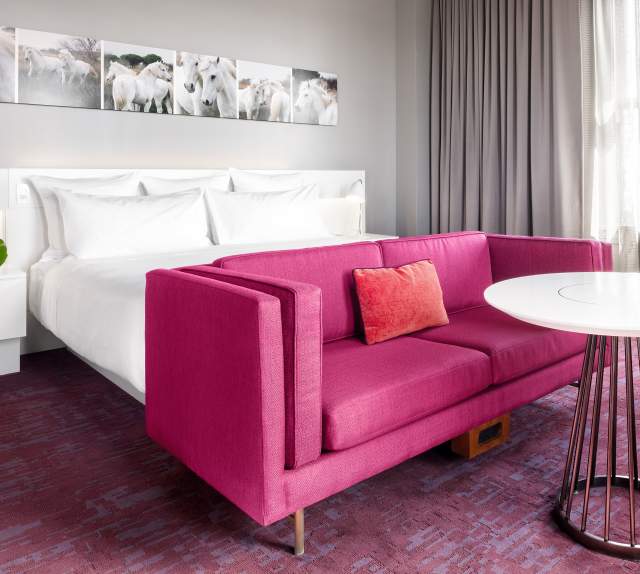 Interior of a very modern and chic room at 21c Museum Hotel. It has a white bed and nightstand, and red couch at the foot of the bed.