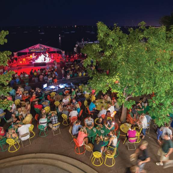 Dozens of people watching a concert at Memorial Union Terrace at night. The photo is taken from above showing the stage, people, and the popular yellow, orange, and green terrace sunburst chairs.