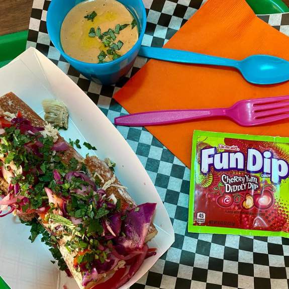 A view from above of an open-face sandwich, a side of yellow soup, Fun Dip and brightly colored cutlery