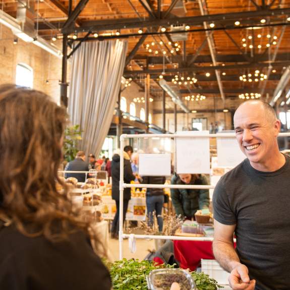 A white man smiling while working a booth at an indoor farmers' market.