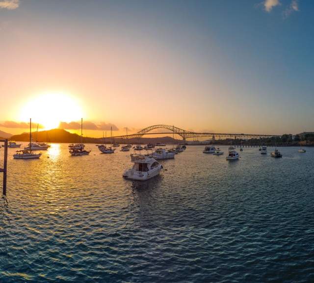 Beautiful sunset over a harbor. The sun is setting behind a bridge of the Americas in the distance, casting a golden light on the water. There are many boats anchored in the harbor, and people can be seen walking along the docks.