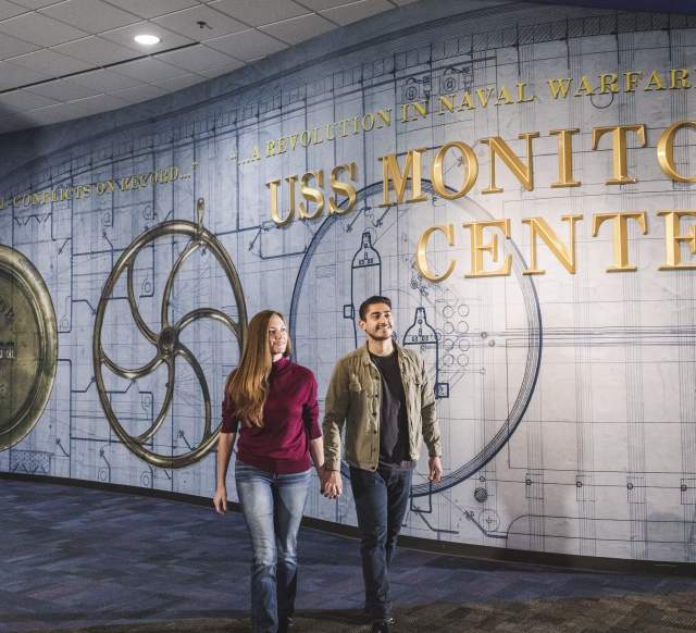 A couple walks in the Mariners Museum, behind them is a sign that reads "USS Monitor Center"