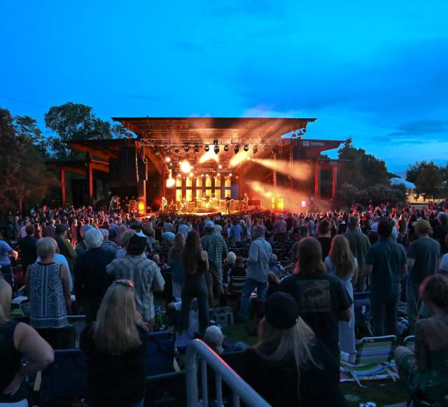 Crowd of people at dusk watching a brightly lit stage with a cool late evening sky
