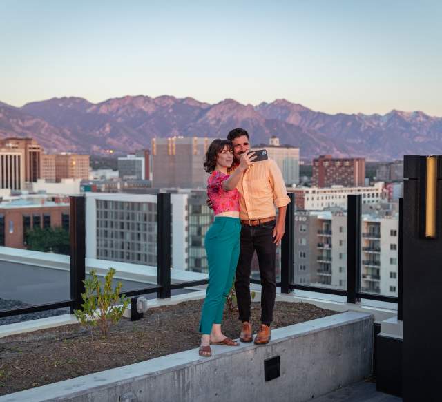 Couple standing and taking a selfie on the roof at Van Ryder with Salt Lake City Skyline and wasatch mountains in the background