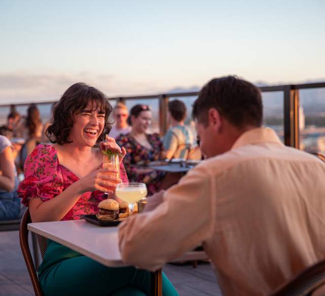 A man and a woman having dinner at a table. The woman is smiling and laughing. The background is a sunset shot over Salt Lake