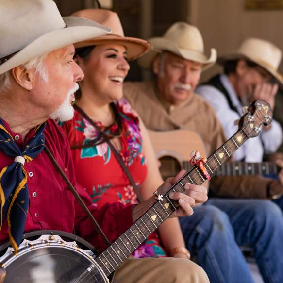 An older man with facial hair sits in the front playing a banjo. He his wearing a red button up shirt, a blue bandana, and a tan cowboy hat. A woman in red, a man playing a guitar, and a man playing harmonica are sitting behind him.
