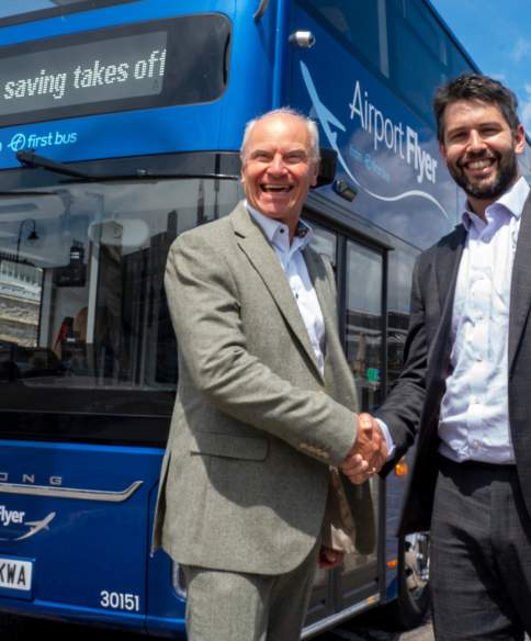 Two men shaking hands in front of a dark blue electric bus parked outside Bristol Temple Meads railway station.