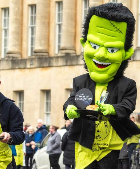 A man dressed in Regency costume and a man dressed in a Frankenstein costume running along Bath's Royal Crescent while holding trays containing a champagne flute and bun.