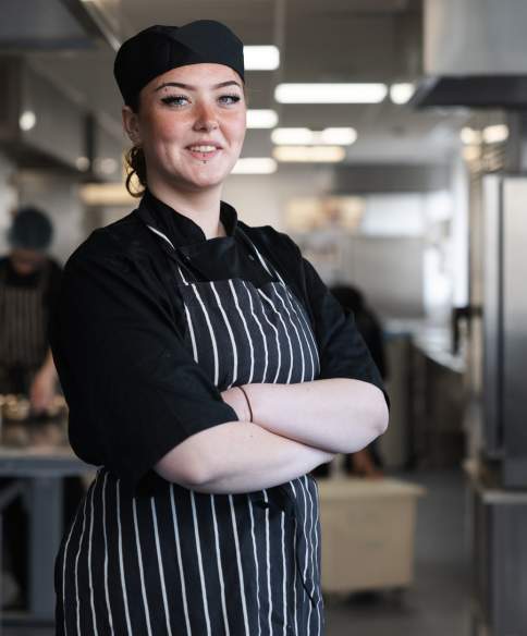 A chef standing with their arms folded and smiling in the centre of a commercial kitchen.