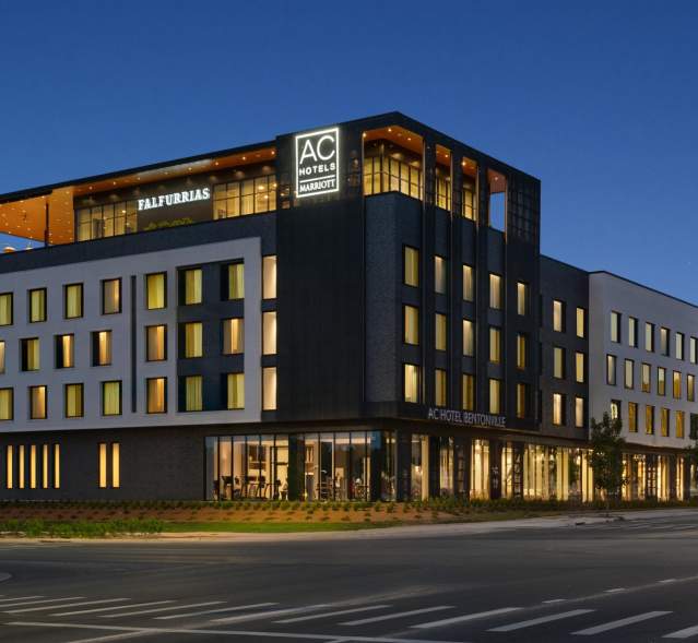 Modern hotel building at dusk, featuring sleek architecture and illuminated signage. Surrounding greenery and street view enhance the inviting atmosphere.