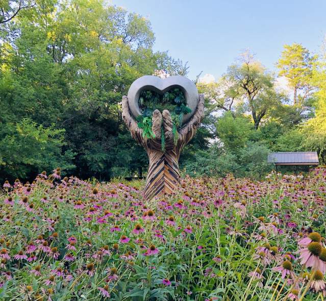 A large sculpture with hands that hold a heart. Pink flowers surround the sculpture.