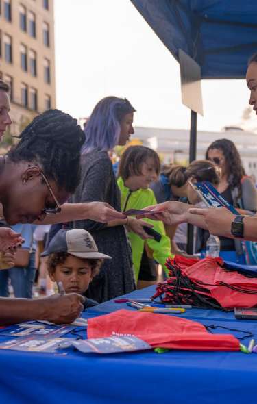 A woman in a bright yellow "Volunteer" shirt hands race bib to a kid and their parent for the IronKids race.