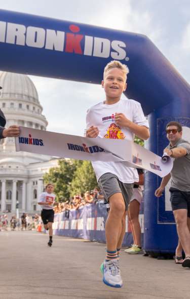 A young white kid runs across the finish line under the Ironkids arch in front of the State Capitol