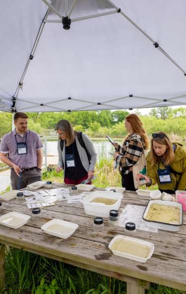 Environmental meeting attendees gather to view specimens under a tent outdoors