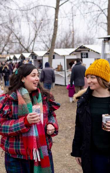 Two women at an outdoor winter market talking and holding drinks.