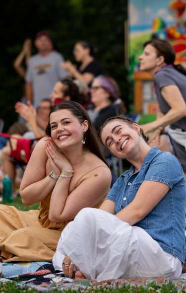Two young women are sitting on blankets in the grass amongst others smiling at the camera while enjoying live music.